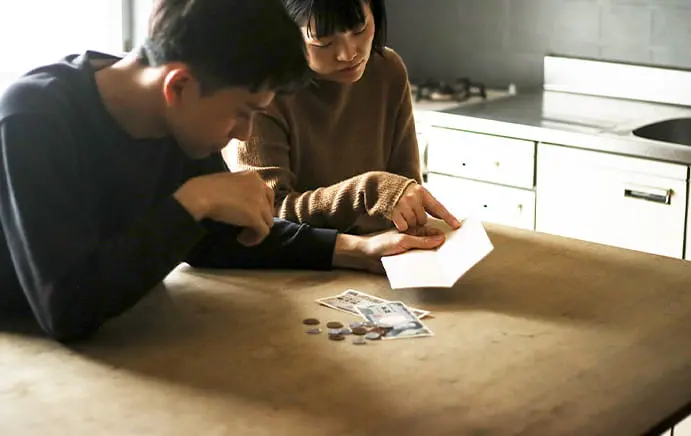 暗いキッチンで若い男女がテーブルに座り、通帳とお金を見ながら真剣な表情で話し合っている