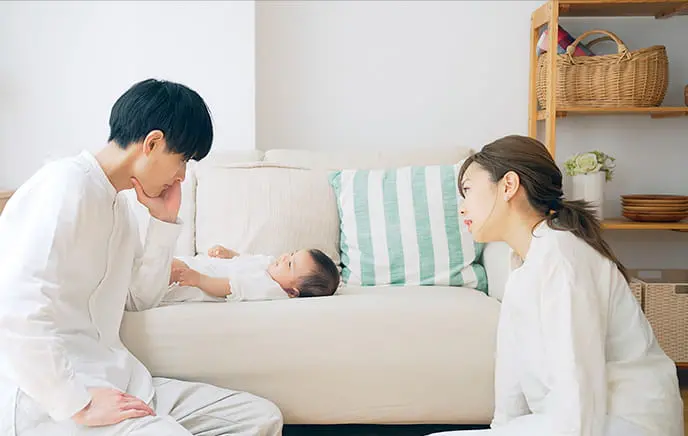明るいリビングで赤ちゃんを囲み、両親が向かい合って座りながら見守っている様子