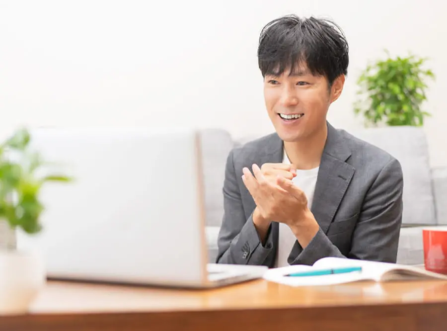 スーツ姿の男性がノートパソコンの前で笑顔を見せながらオンラインミーティングをしている様子